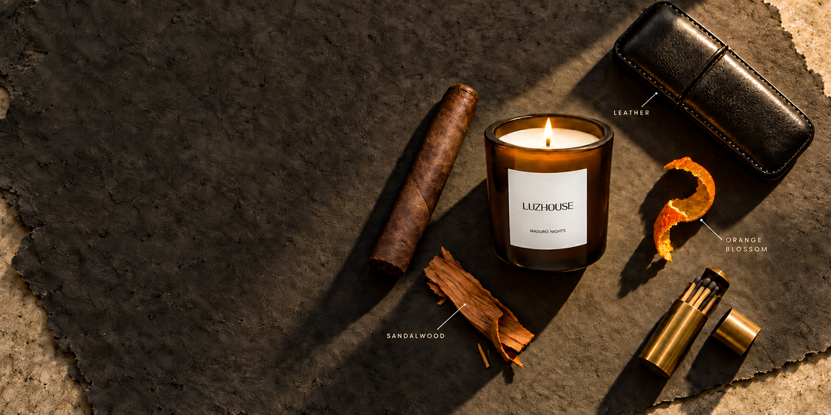 A luxury candle in a dark amber glass jar with a white label reading 'LUZHOUSE', styled on a dark textured surface with a cigar, sandalwood piece, orange peel, a black leather case, and a brass lighter.
